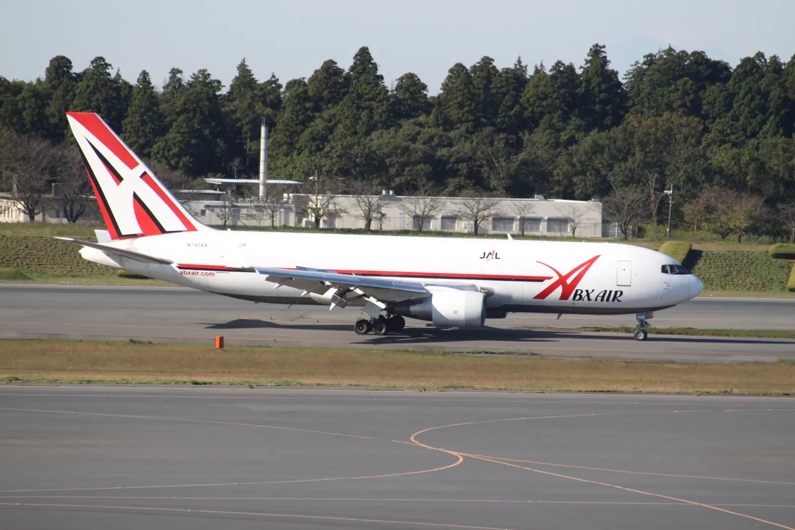 ABX Air Boeing 767F cargo plane on a runway, with a red, white, and black livery, trees and buildings in the background.