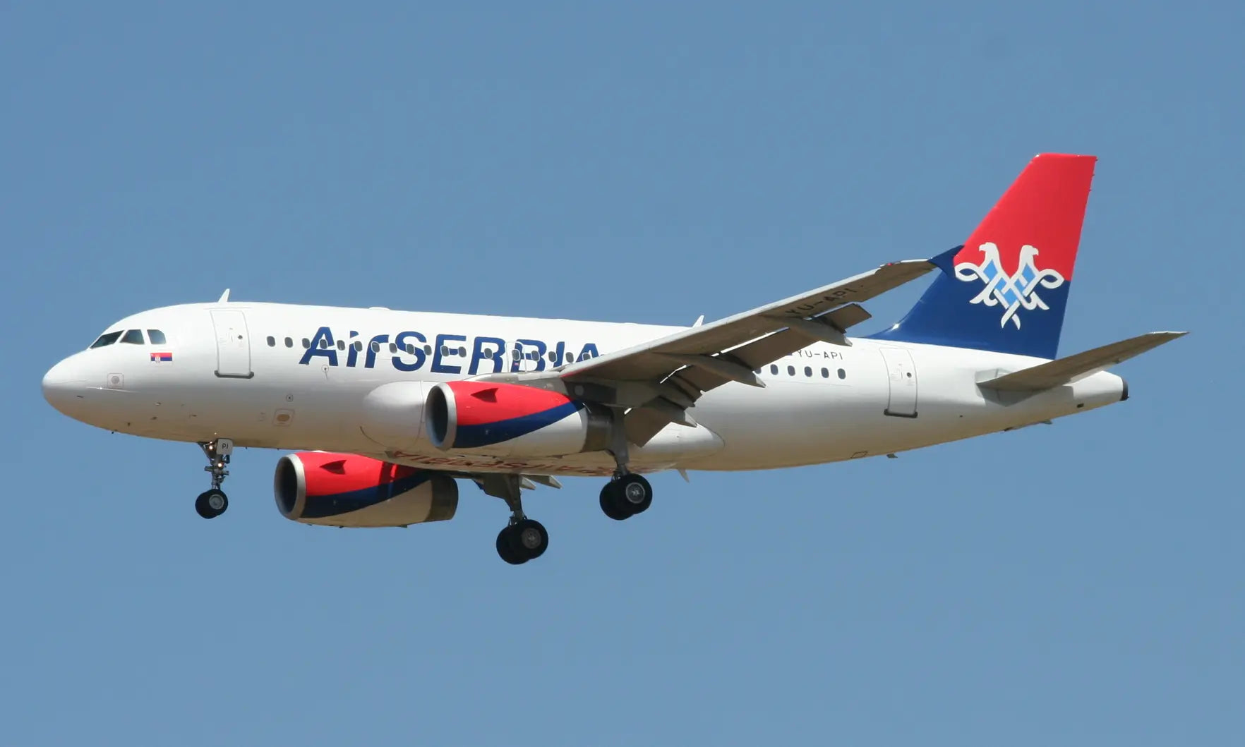 Air Serbia plane in flight with landing gear deployed against a clear blue sky.