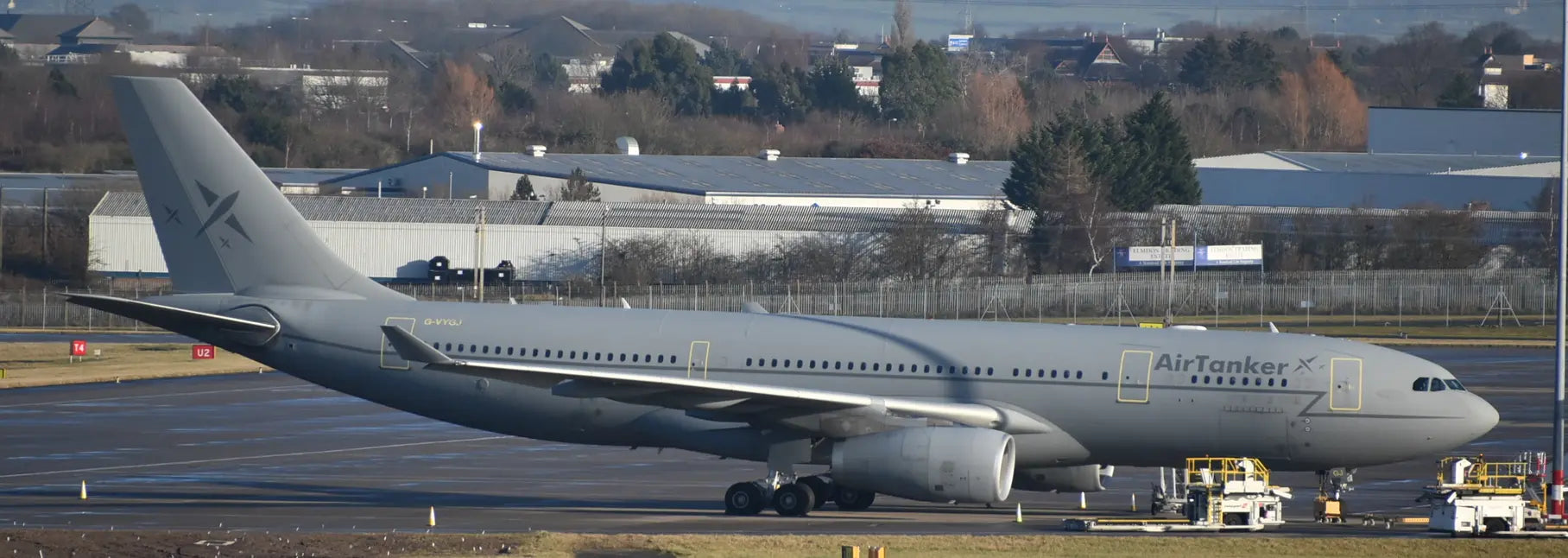 AirTanker Holdings Ltd. Airbus A330-243 aircraft parked on tarmac at Birmingham Airport, with industrial buildings and trees in the background.