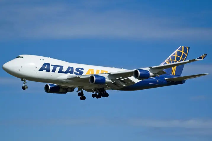 Atlas Air Boeing 747-400F cargo plane in mid-flight, with landing gear deployed, against a clear blue sky.