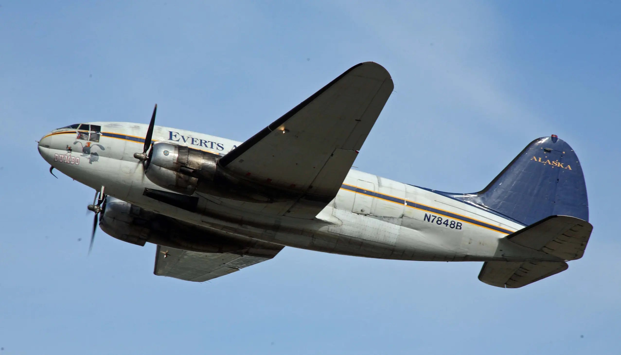 Everts Air Cargo C-46 Commando aircraft taking off with visible propellers, labeled 'Dumbo,' against a clear blue sky.