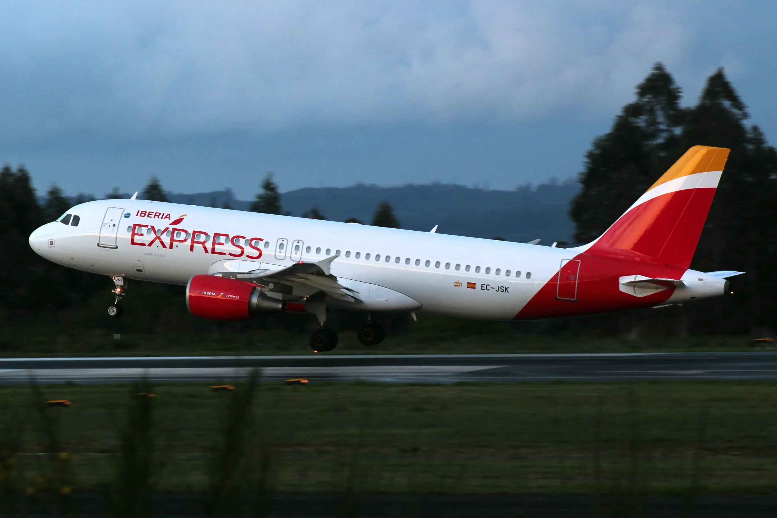 Iberia Express Airbus A320 landing on a runway with landing gear extended, set against a cloudy sky and distant trees.