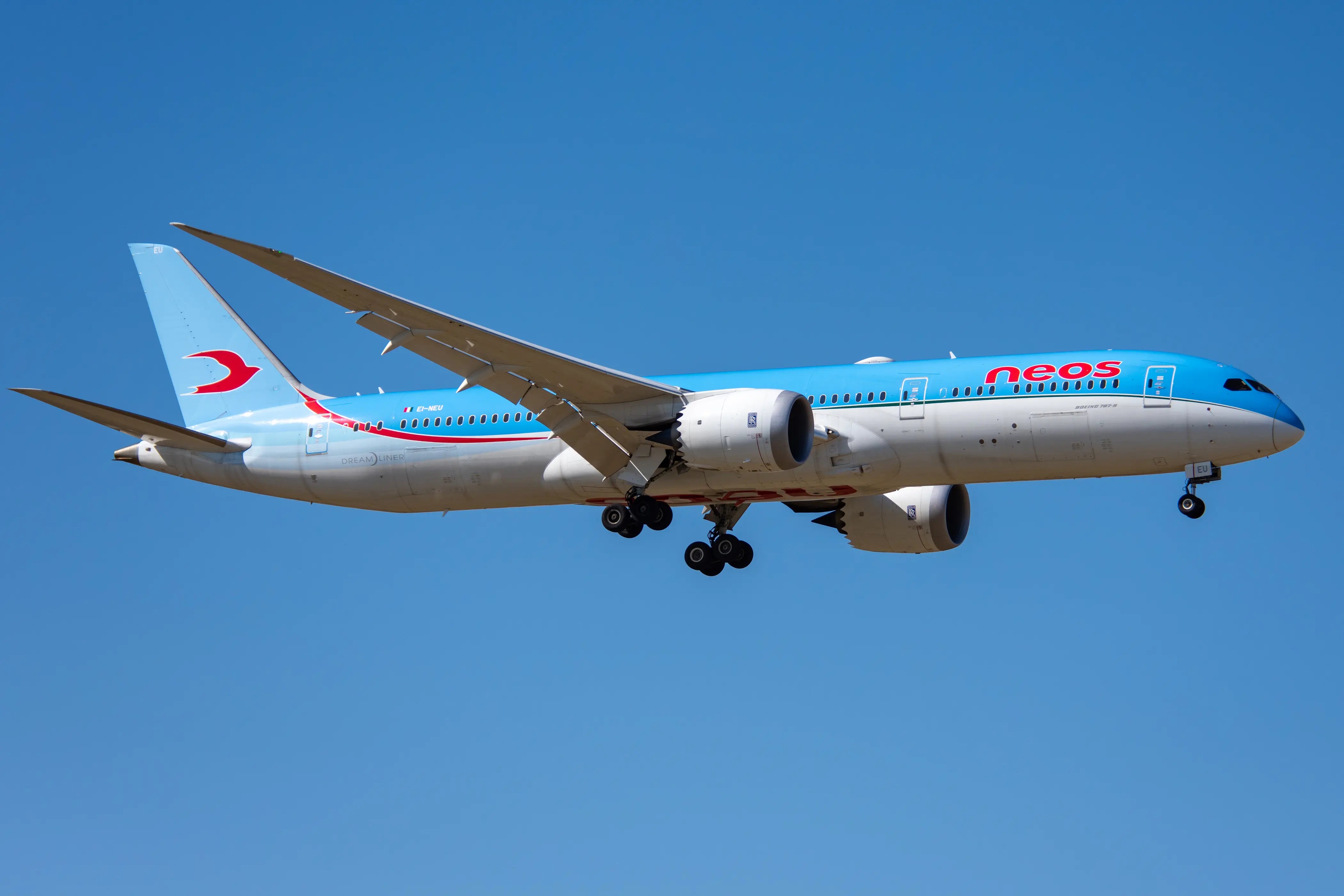 A Neos Boeing 787 Dreamliner in flight, against a clear blue sky, with landing gear extended.