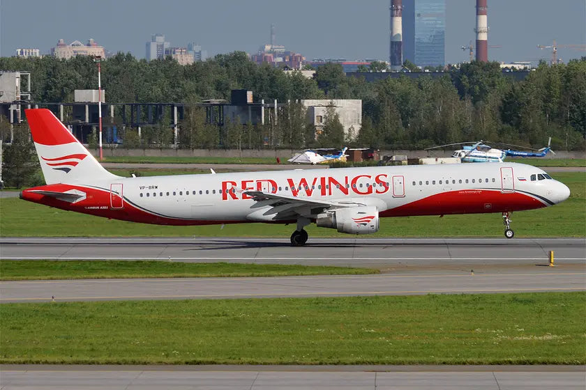 Side view of a Red Wings Airbus A321-211 aircraft parked on a runway, with green grass and buildings in the background.