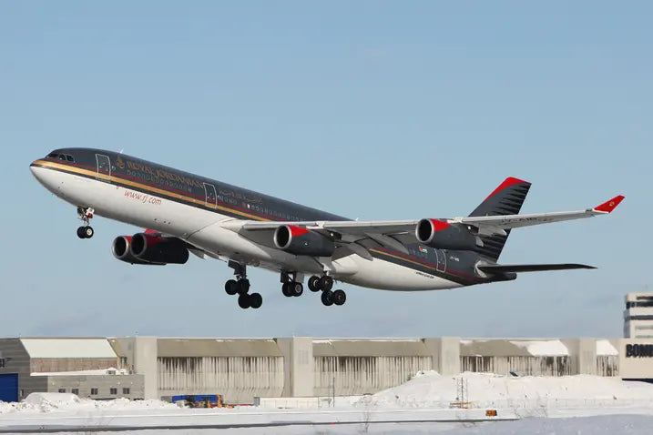 Royal Jordanian Airbus A340 taking off against a clear blue sky, with snowy ground and buildings in the background.