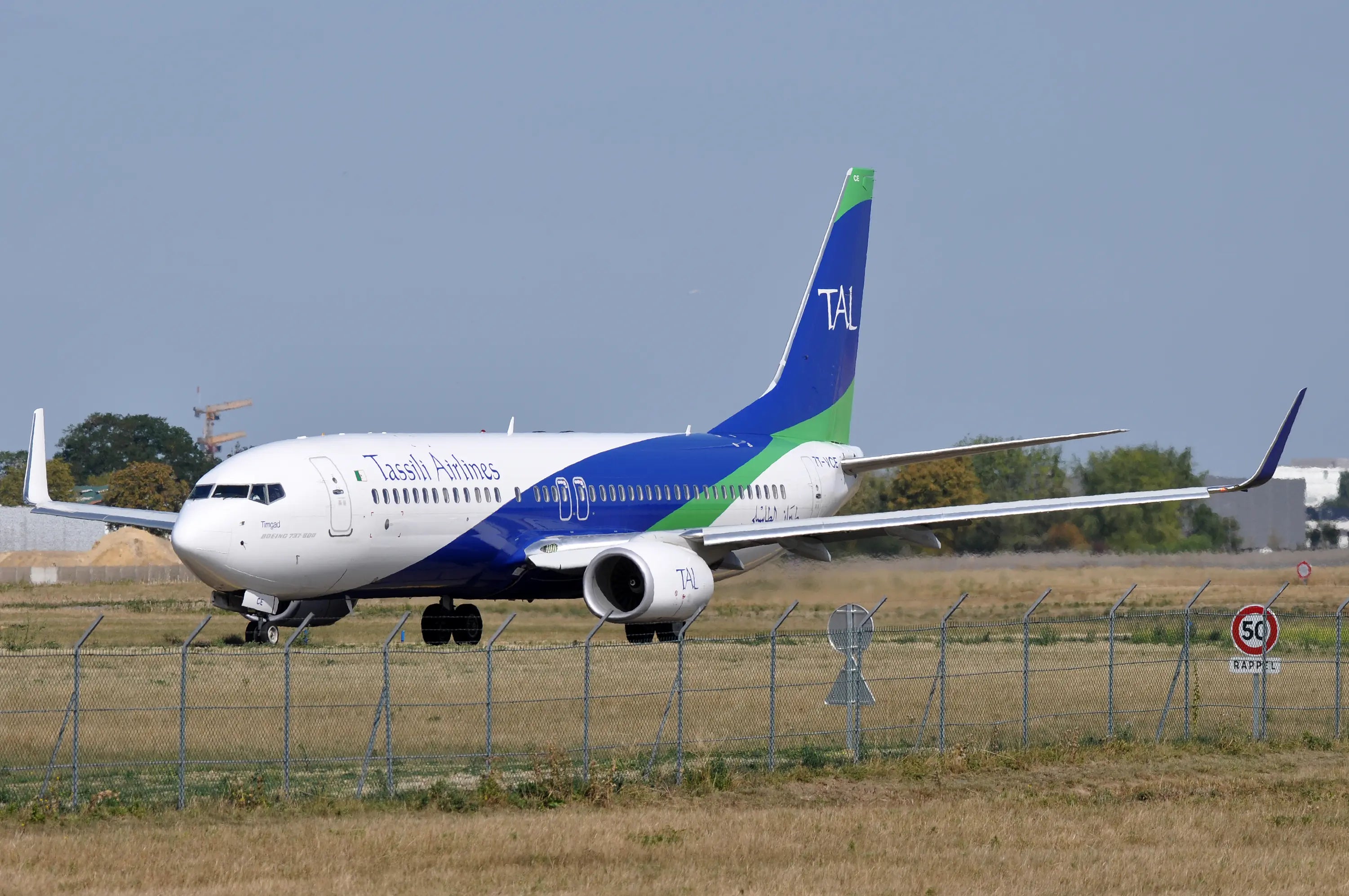 Tassili Airlines Boeing 737-800 on the runway, with distinctive blue and green tail, at an airport on a clear day.