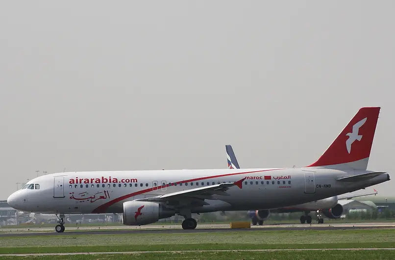 Air Arabia Airbus A320 parked on airport tarmac with visible airline branding and red tail fin, preparing for departure.