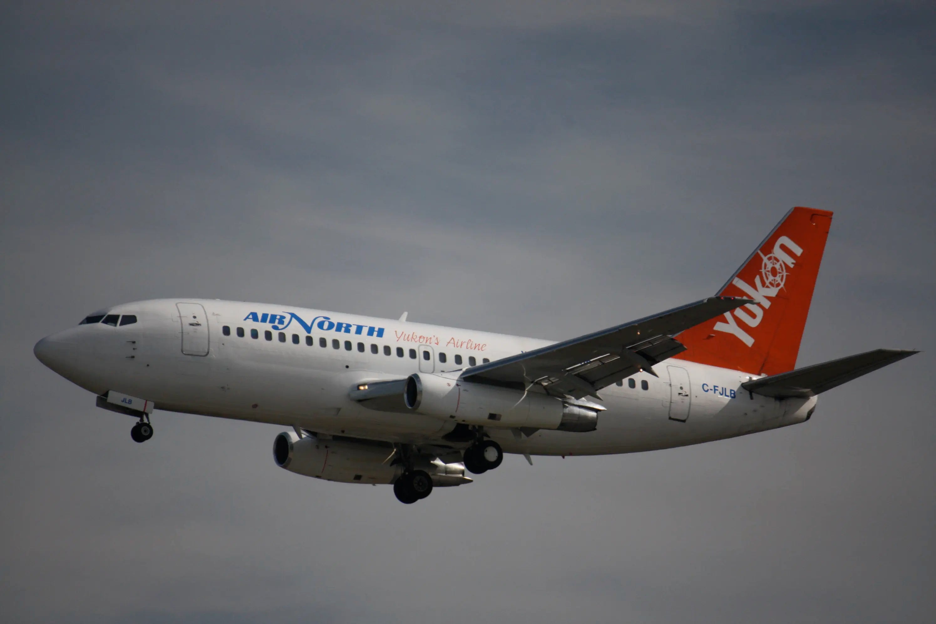 Air North airplane with Yukon logo on tail, flying against a cloudy sky, with landing gear extended.