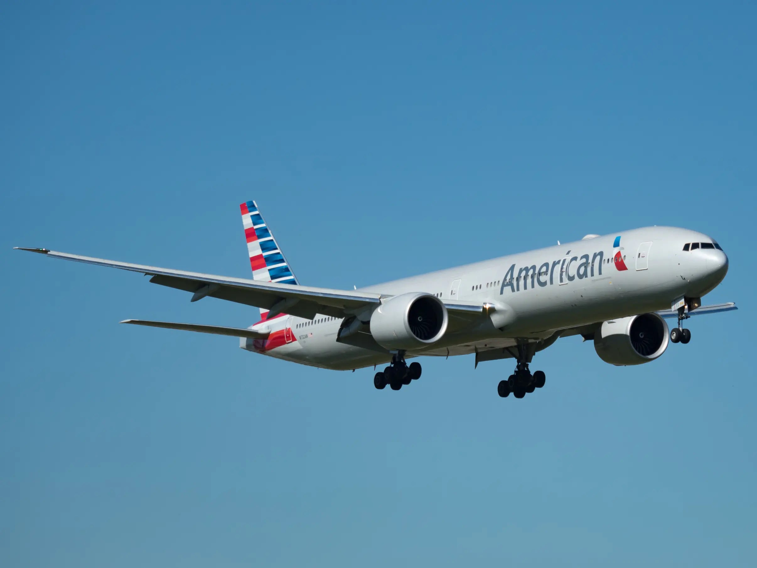 American Airlines Boeing 777-300ER aircraft in flight with landing gear down at Miami International Airport against a clear blue sky.