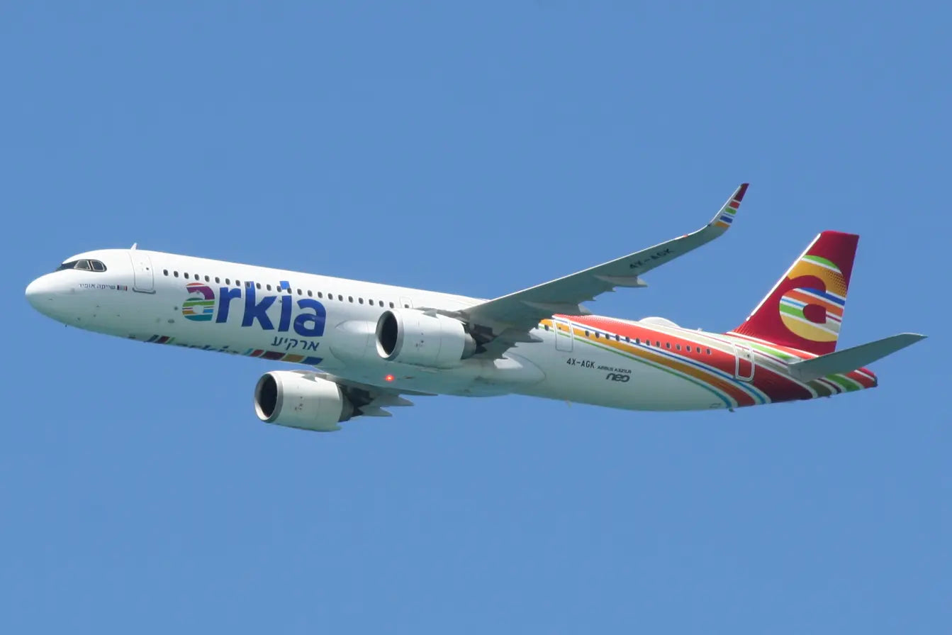 A white Arkia passenger airplane in flight against a clear blue sky, showcasing colorful branding and airplane model 4X-AGK Airbus A321neo.