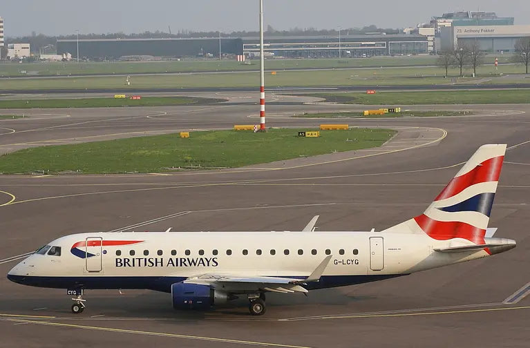 British Airways Embraer E-170 aircraft on a runway at an airport, with terminal building and taxiways visible in the background.