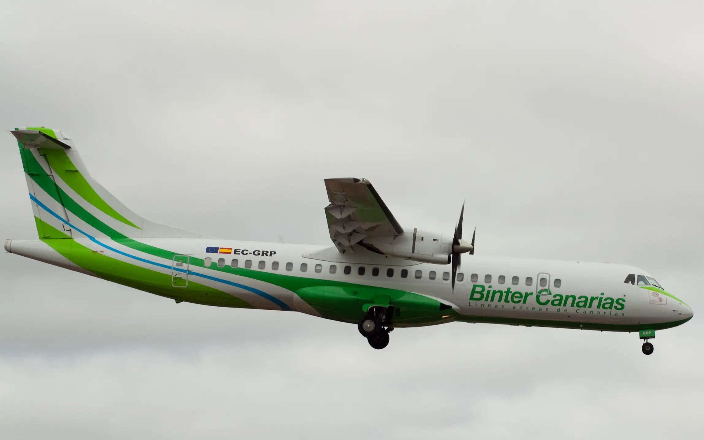 A Binter Canarias ATR 72 aircraft in flight, with green and white livery and registration EC-GRP, against a cloudy sky background.