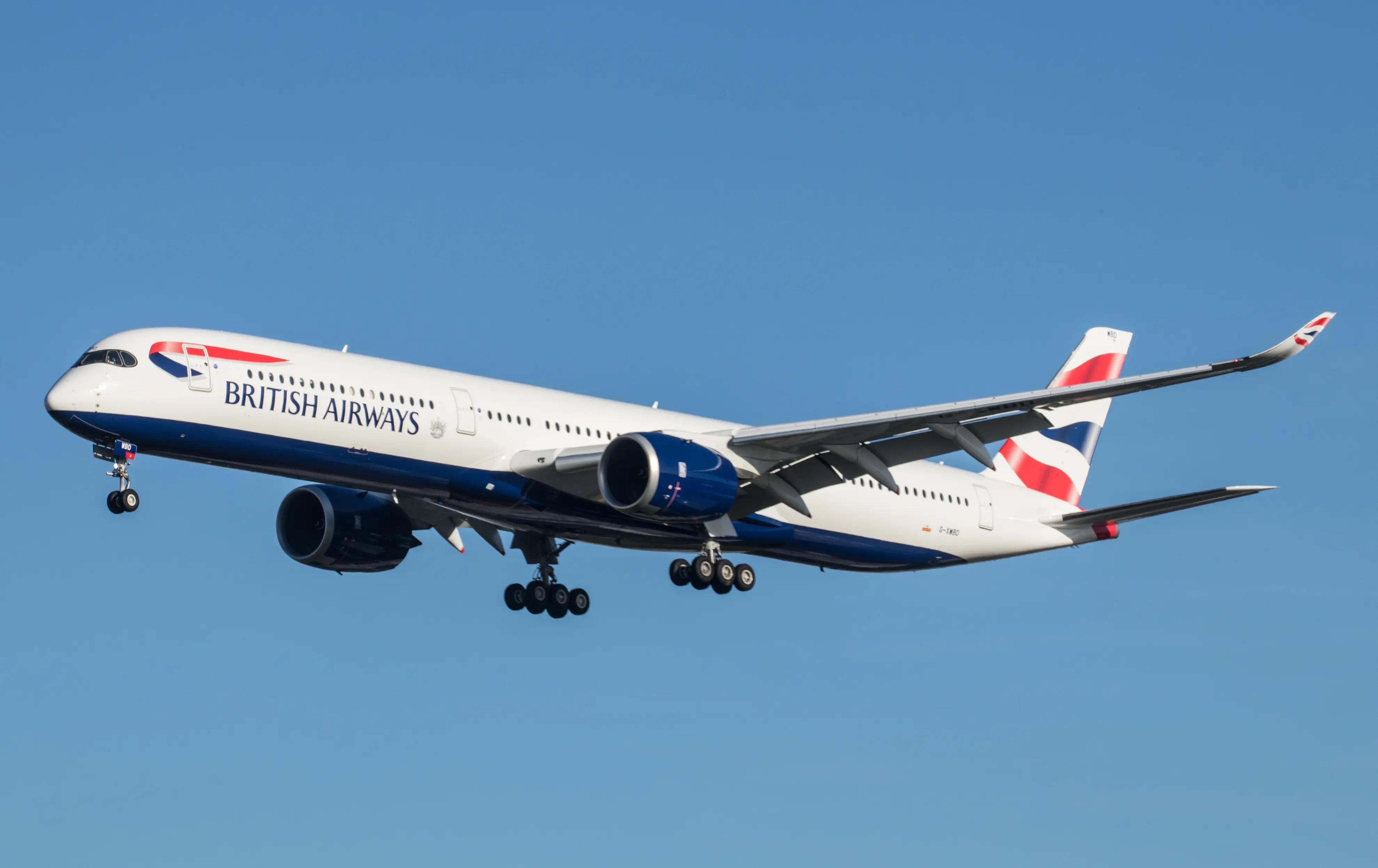 British Airways A350-1000 aircraft in flight against a clear blue sky, with landing gear visible, approaching landing at London Heathrow Airport.