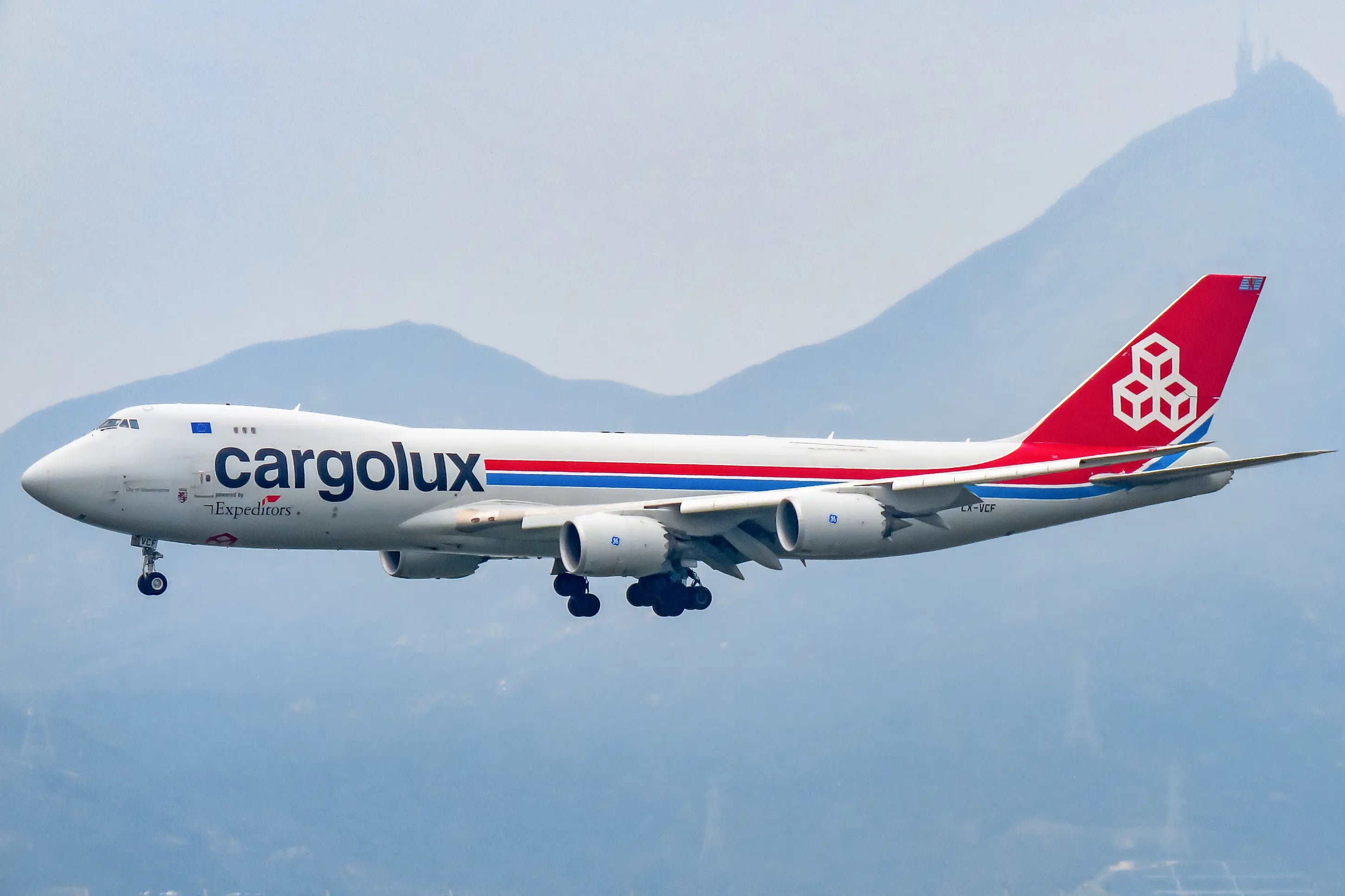 A Cargolux Boeing 747 cargo plane in flight with landing gear extended, set against a mountainous backdrop at Hong Kong airport.