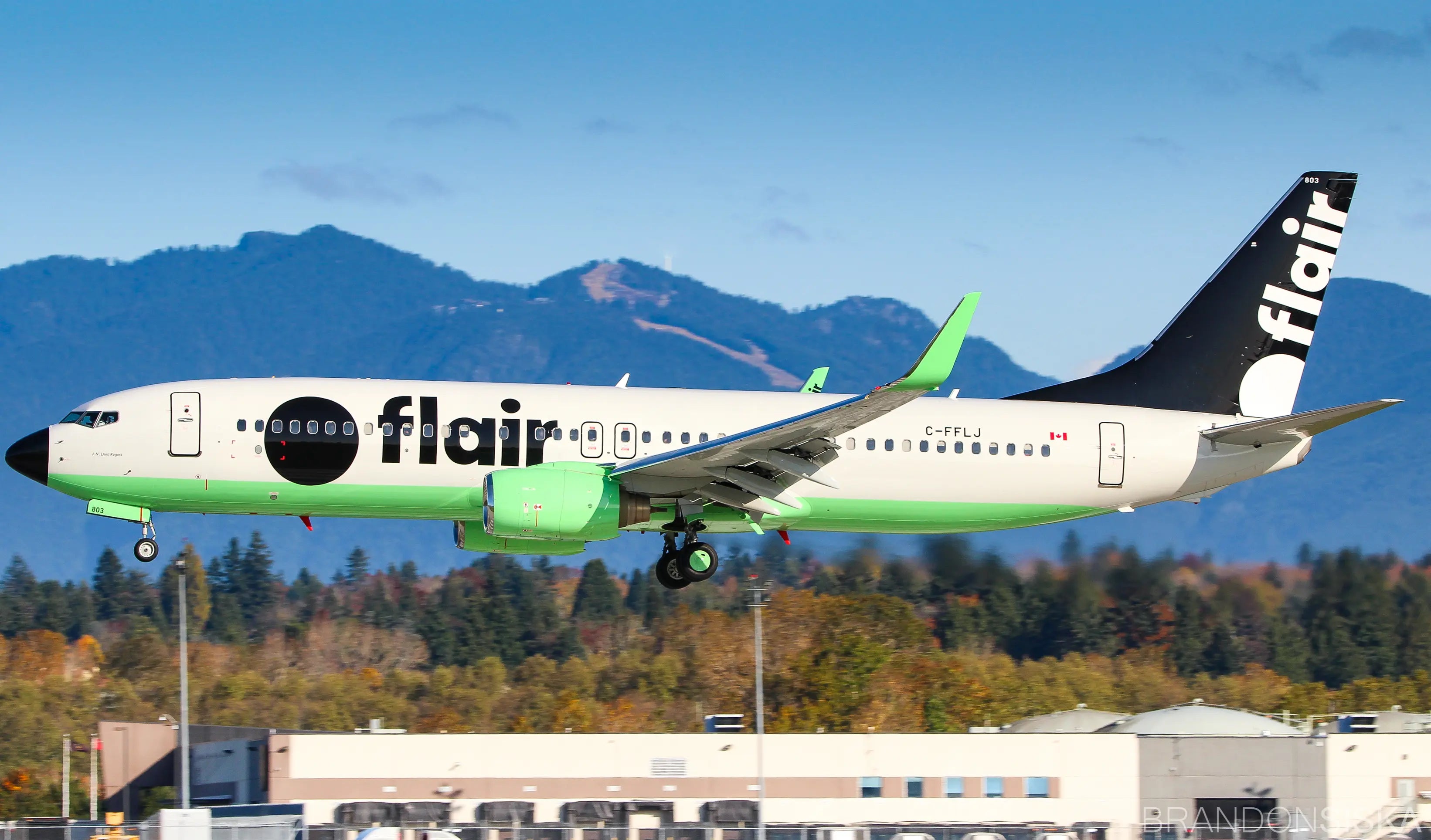 A Flair Airlines plane, registration C-FFLJ, landing at an airport with mountains in the background, its fuselage painted white and green.