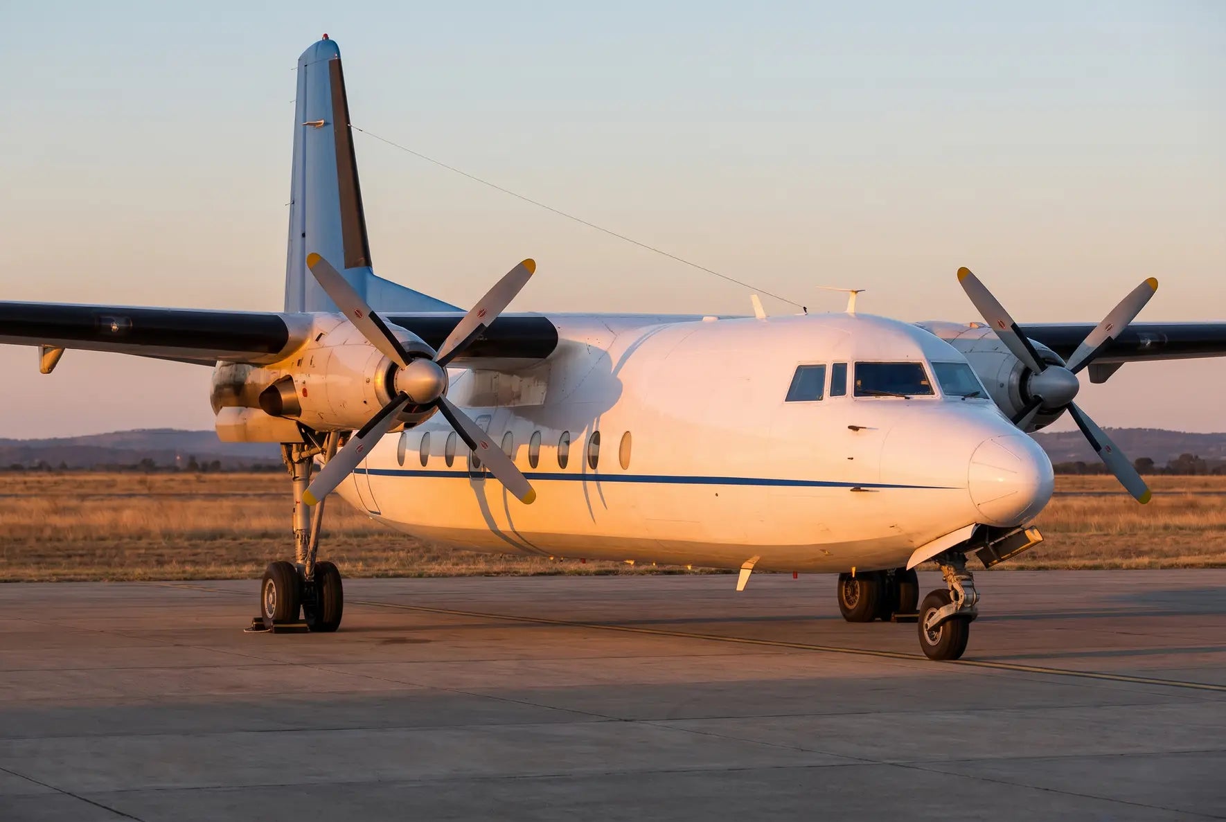 Fokker F27-200 aircraft parked on a runway during sunset with visible propellers and a distant landscape background.