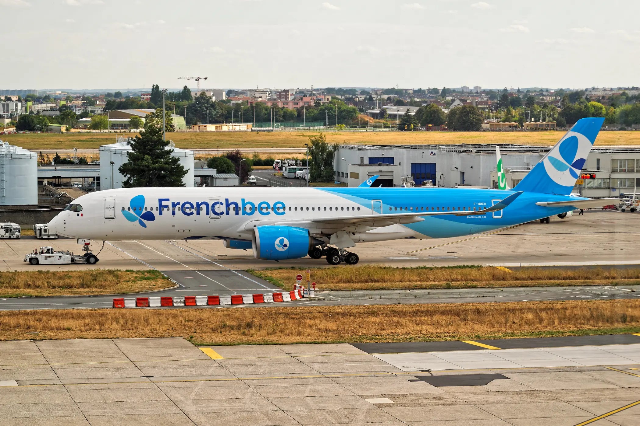 French Bee Airbus A350-941 aircraft parked at an airport with a tow vehicle in front, featuring blue and white livery against an urban landscape backdrop.