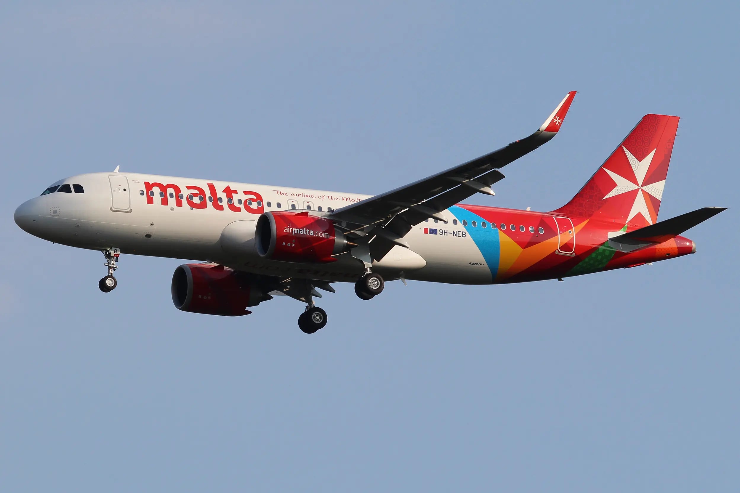 Air Malta Airbus A320neo with registration 9H-NEB in flight, featuring red and white livery and logo, against a clear blue sky.