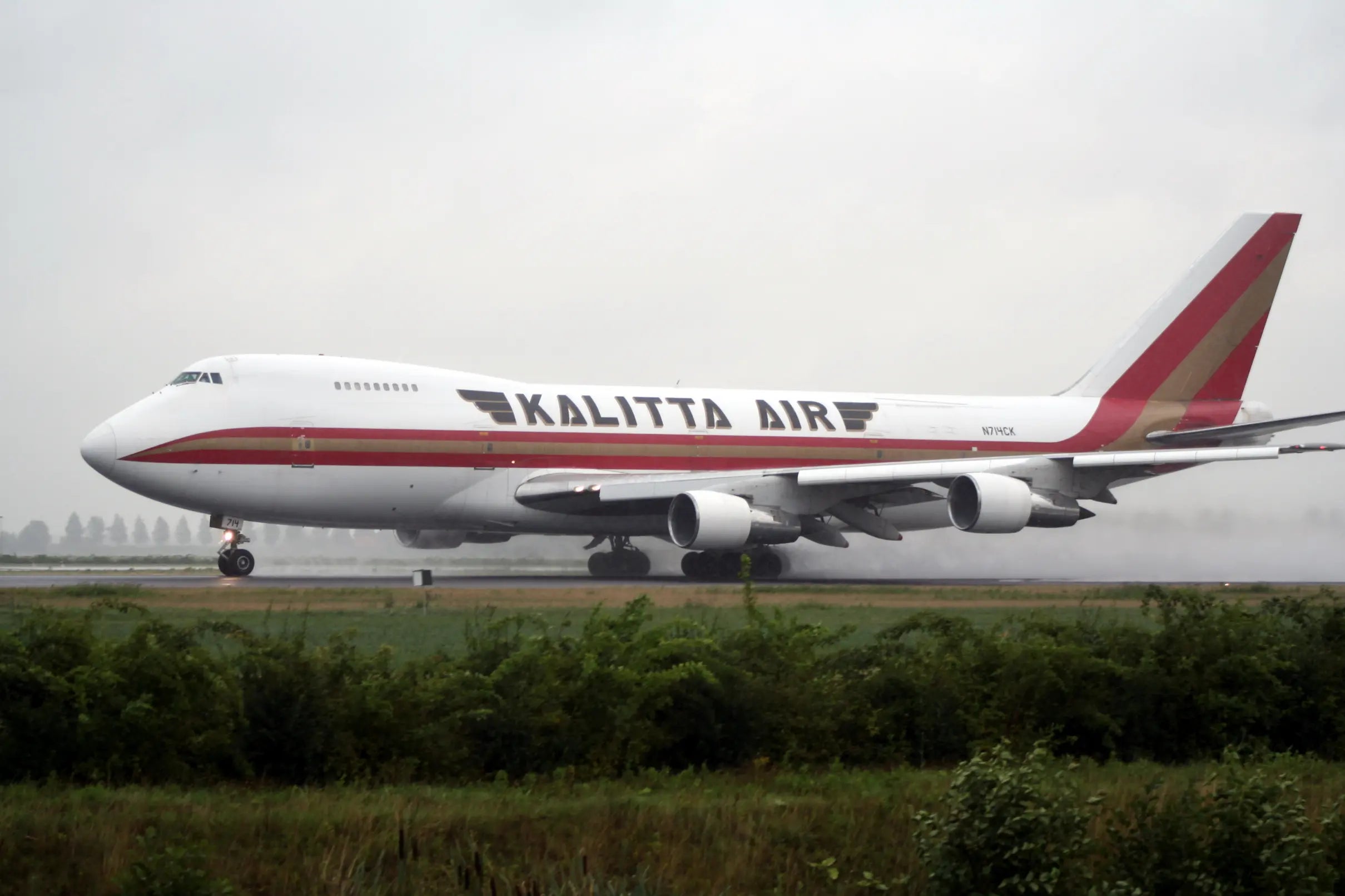 Kalitta Air cargo plane taking off from a wet runway at Schiphol Airport with mist trailing from the wheels, overcast sky in the background.