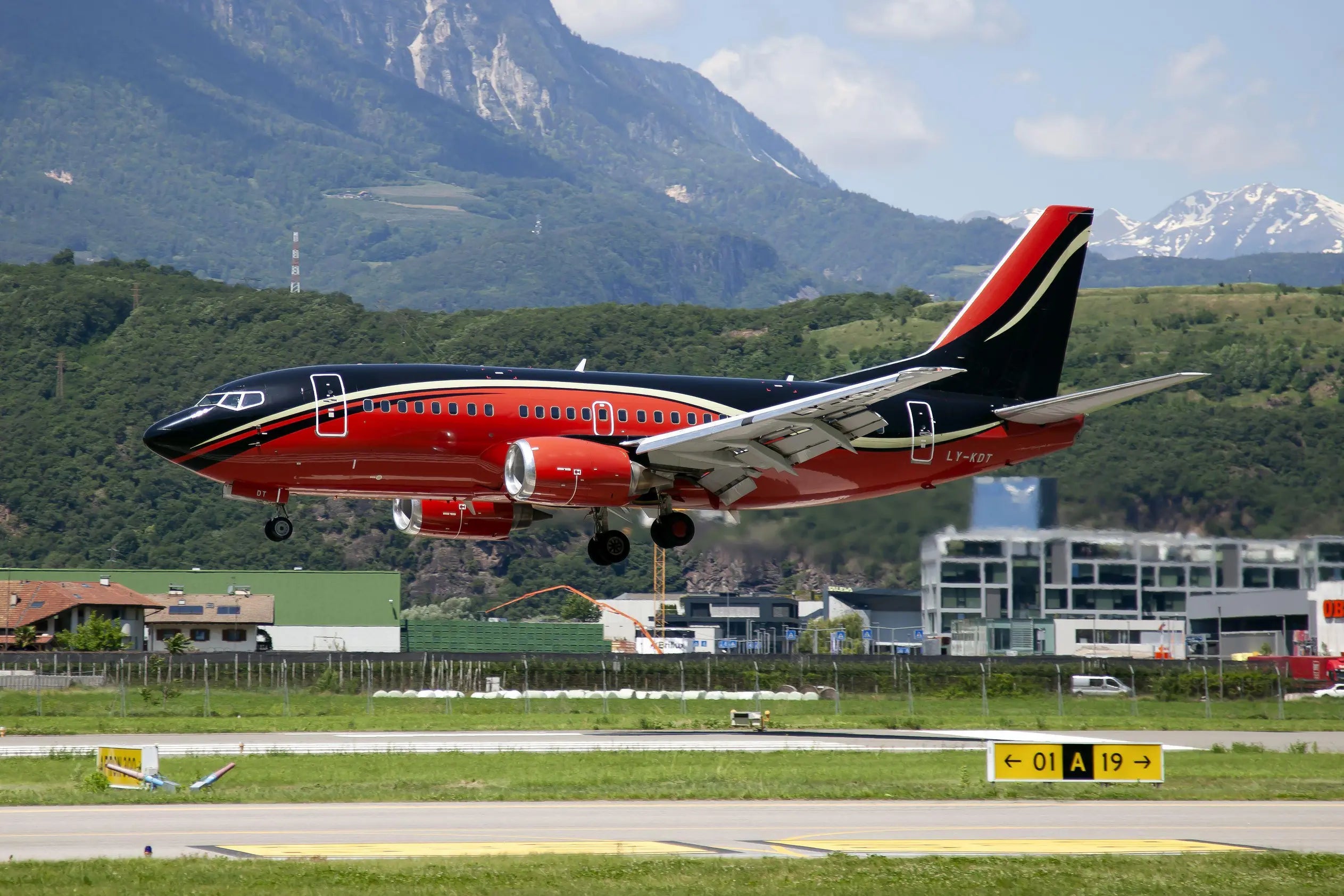 A Boeing 737-500 with tail number LY-KDT taking off from an airport with green hills and mountains in the background.