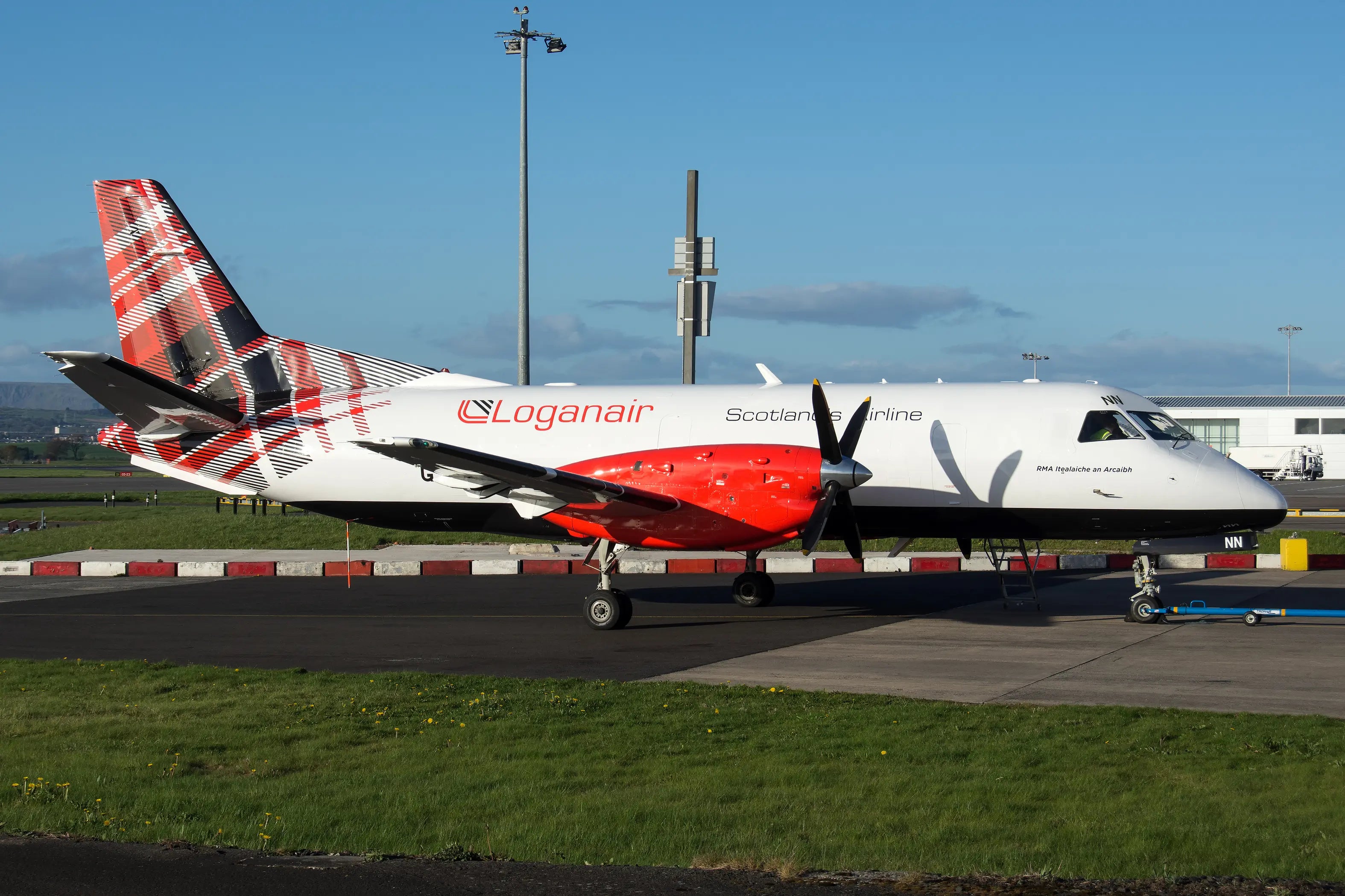 Loganair Saab 340 aircraft with new livery, showcasing a red tartan design on the tail, parked at an airport with clear blue skies.