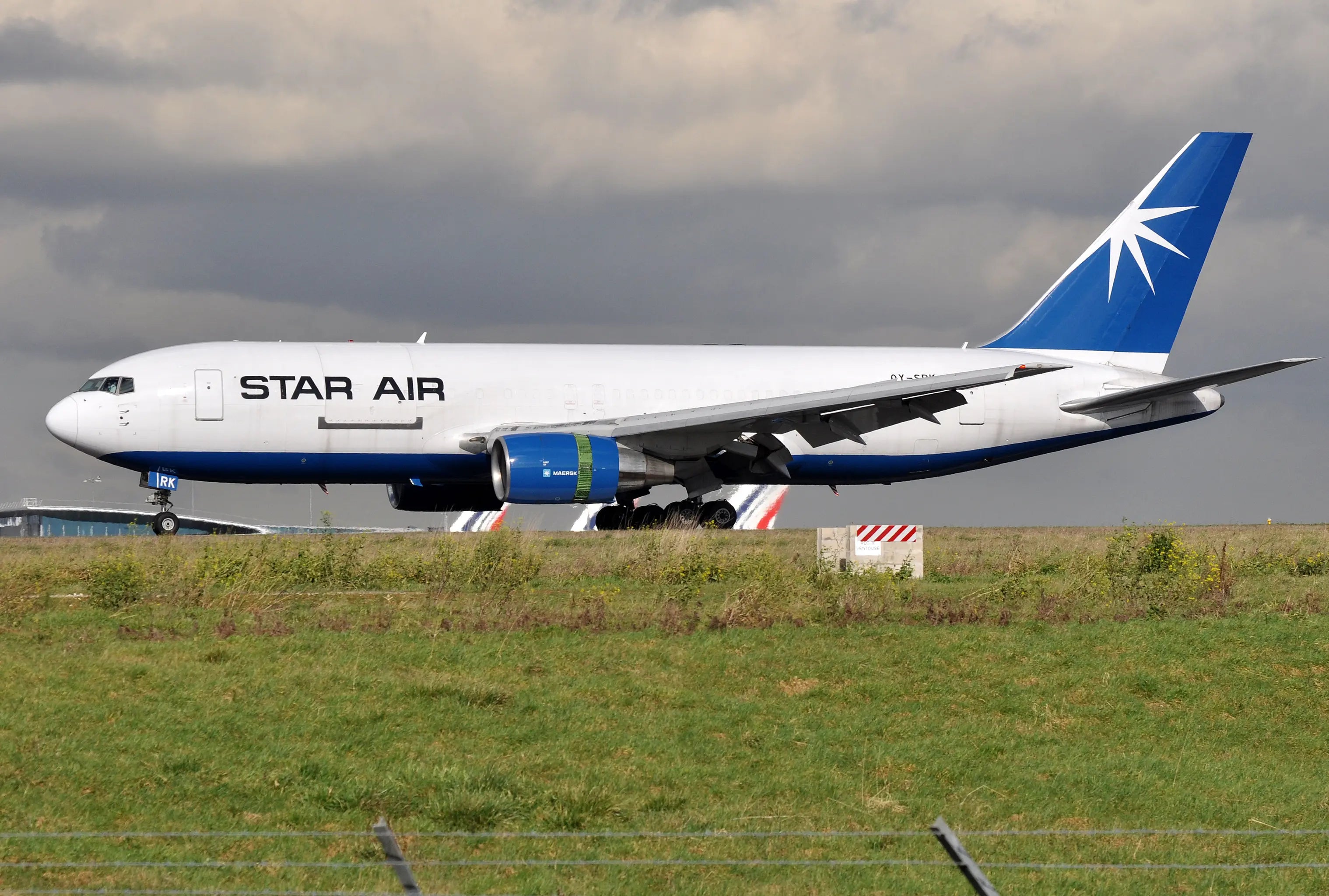 Star Air Boeing 767-204(BDSF) aircraft OY-SRK taxiing on a runway with a grassy field in the foreground and cloudy sky backdrop.