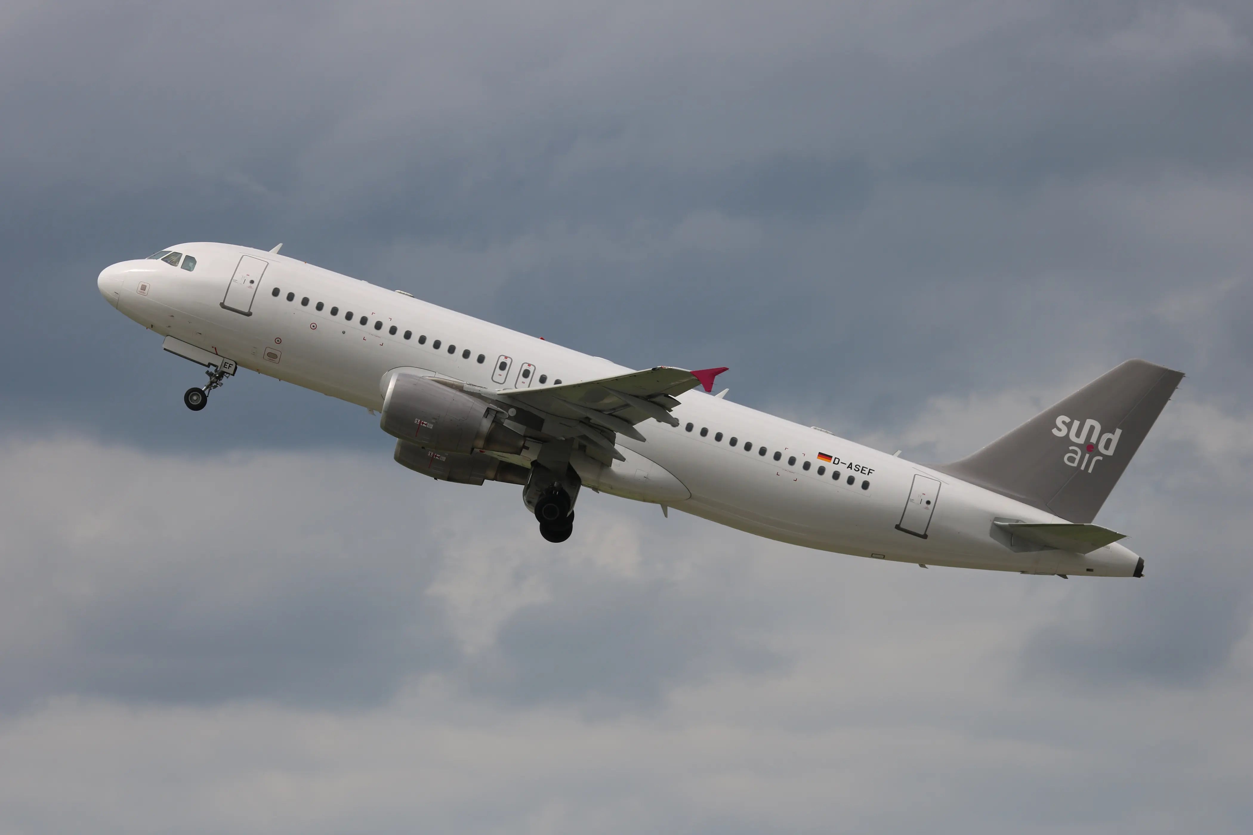 Sundair Airbus A320-200 with registration D-ASEF taking off under a cloudy sky, showing landing gear and airline logo on the tail.