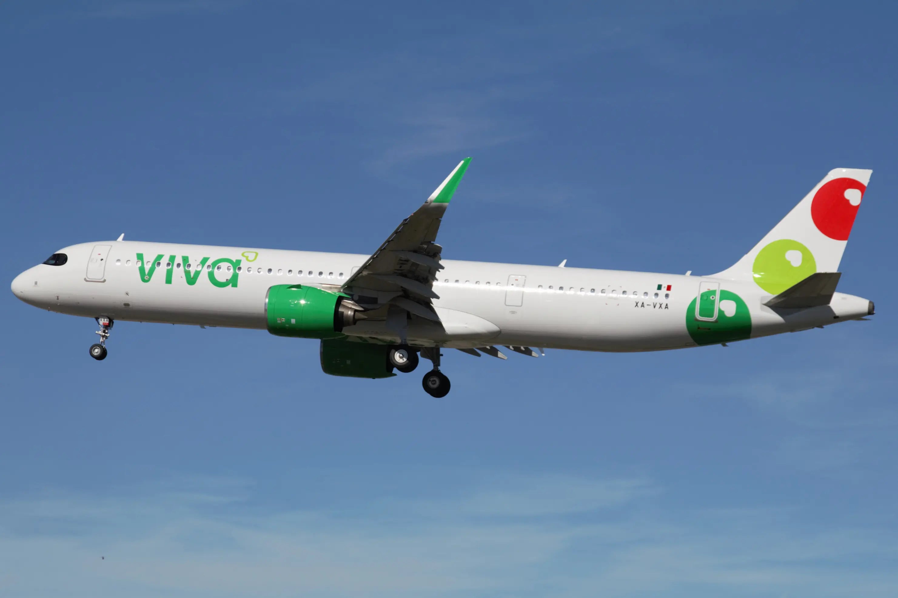 Side view of a Viva Aerobus aircraft in flight against a clear blue sky, featuring green engines and a tail with red and green circles.