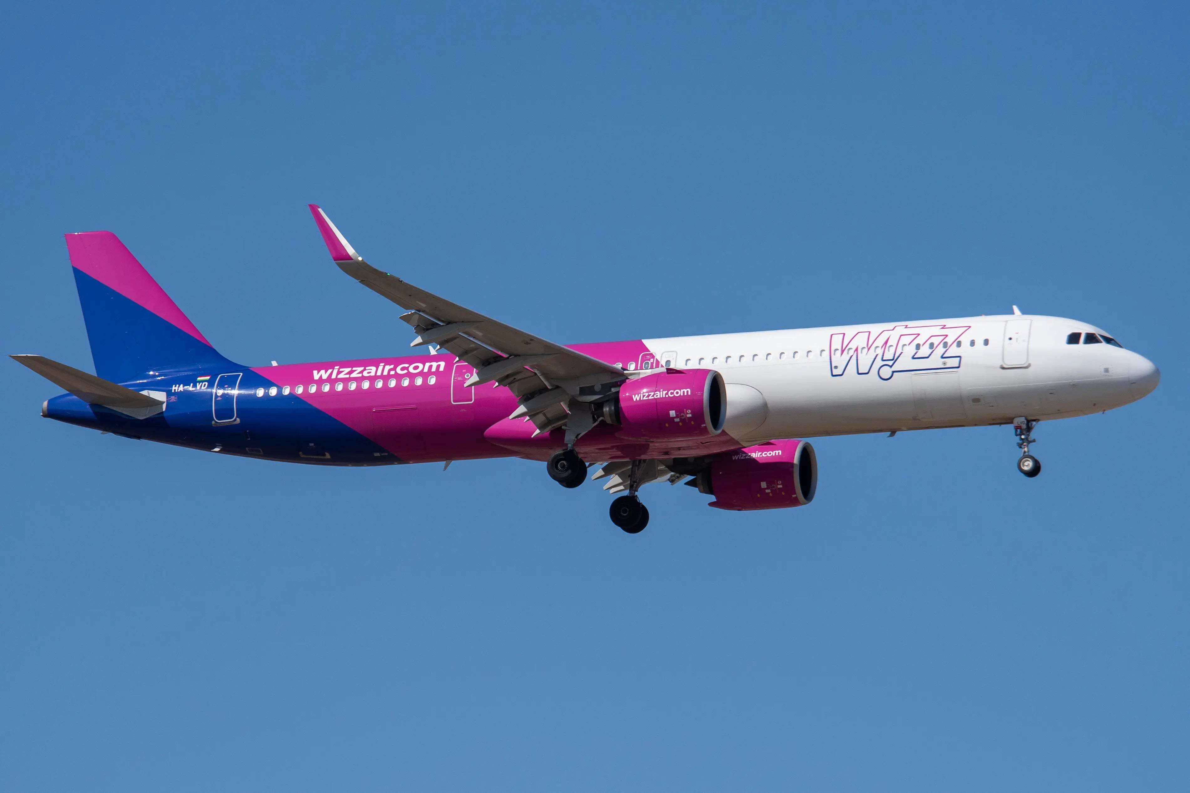 Wizz Air airplane with pink and blue livery flying in clear blue sky, showing landing gear extended.