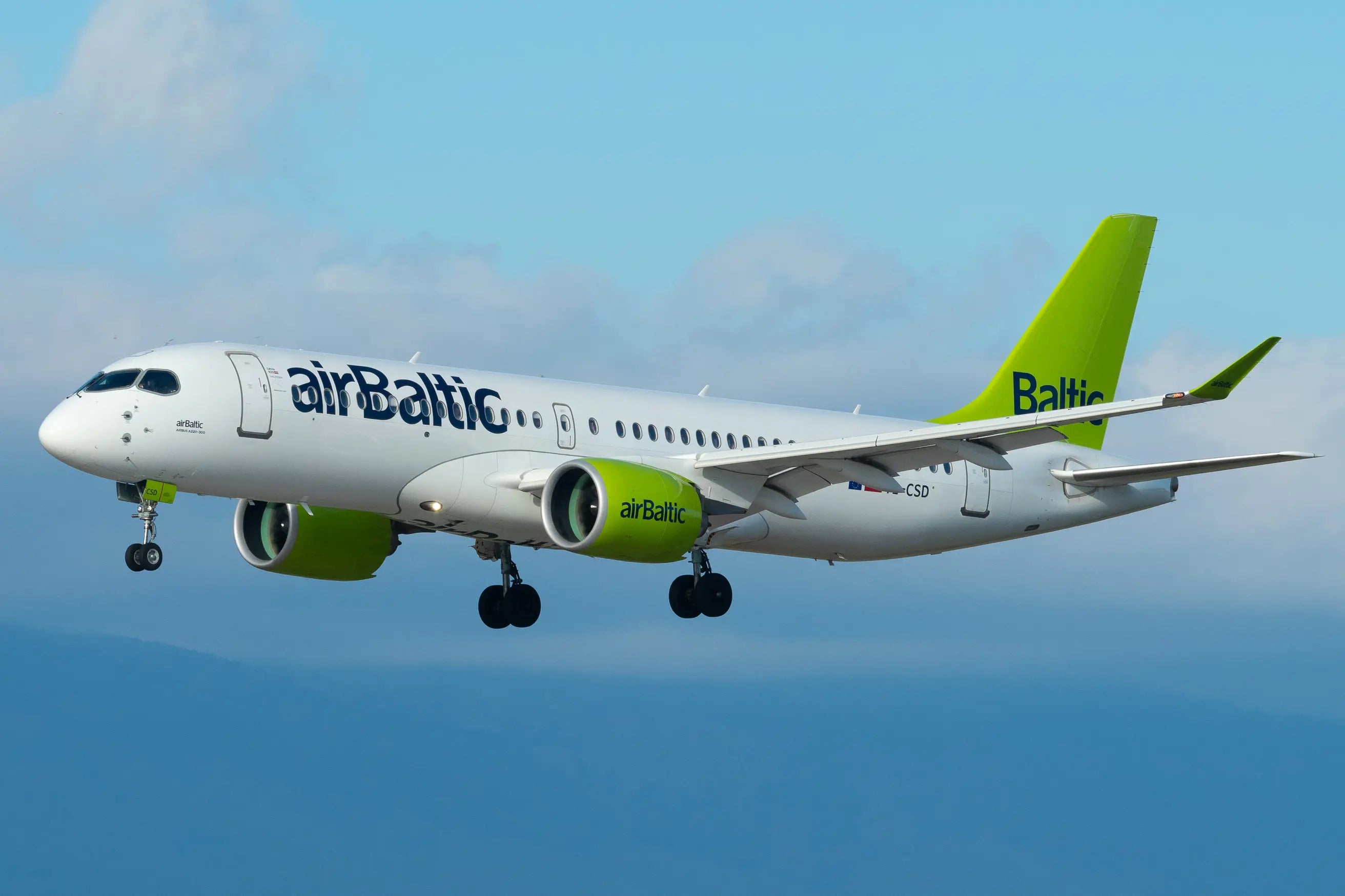 AirBaltic Airbus A220-300 airplane in flight against a blue sky with landing gear extended.